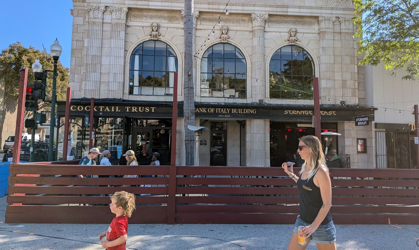 A sunlit street scene on Main Street in Ventura, California shows a historic stone building with arched windows housing bars and restaurants, as a woman holding a drink and a young child walk past an outdoor patio in the foreground.
