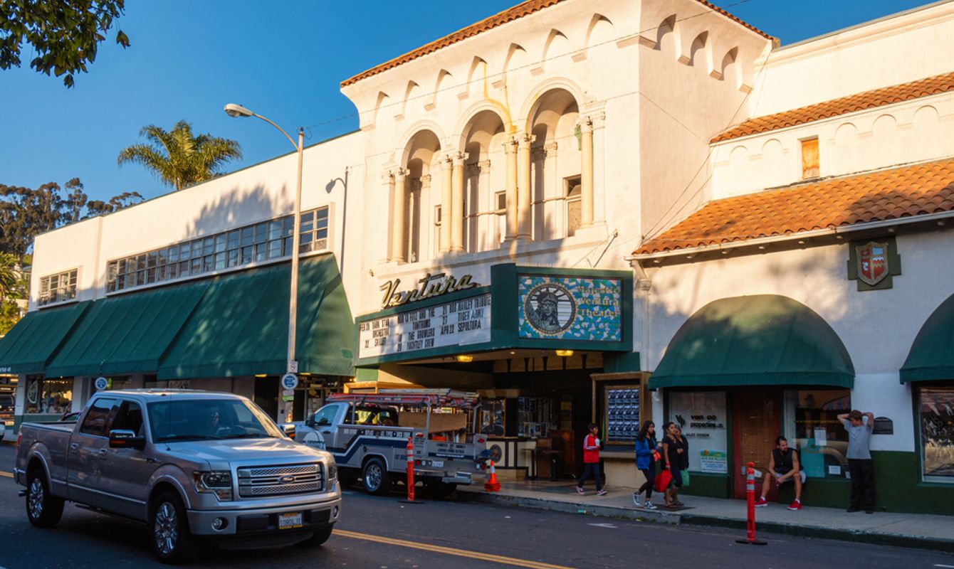 The Ventura Theatre’s Spanish-style facade with arched windows and a marquee sign faces a busy street with parked trucks and pedestrians walking along the sidewalk.
